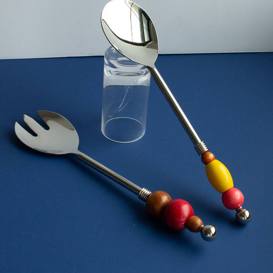 A set of stainless steel salad serving utensils, including a fork and a spoon, with colorful wooden bead handles in red, brown, yellow, and silver, arranged on a blue surface with a clear glass tumbler in the background.