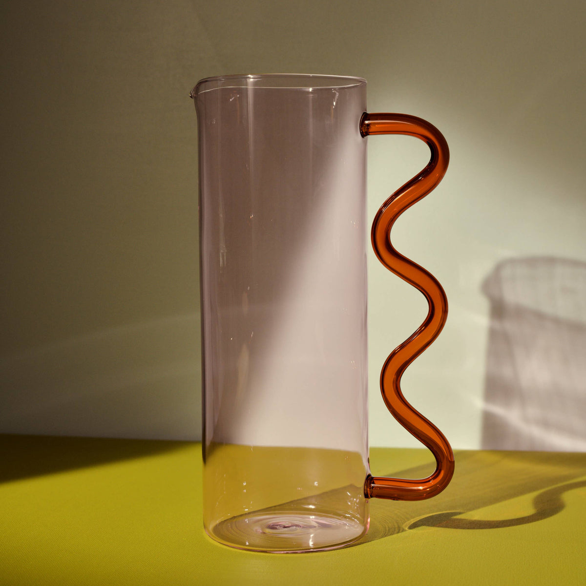 Clear glass pitcher with a wavy brown handle on a yellow surface.