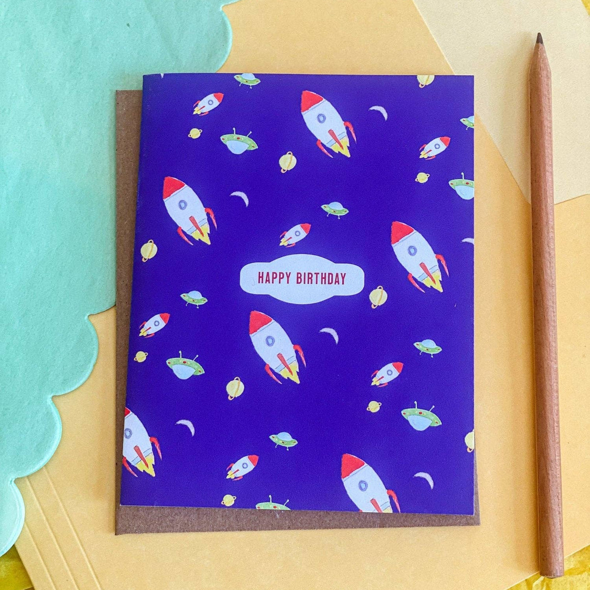 A purple birthday card with a space theme, featuring illustrations of rockets, planets, and stars, with a 'HAPPY BIRTHDAY' label in the center. The card is displayed on a yellow paper background next to a brown pencil and a teal scalloped paper cutout.