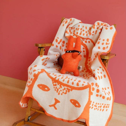 Orange and white patterned blanket draped over a wooden chair against a pink wall.