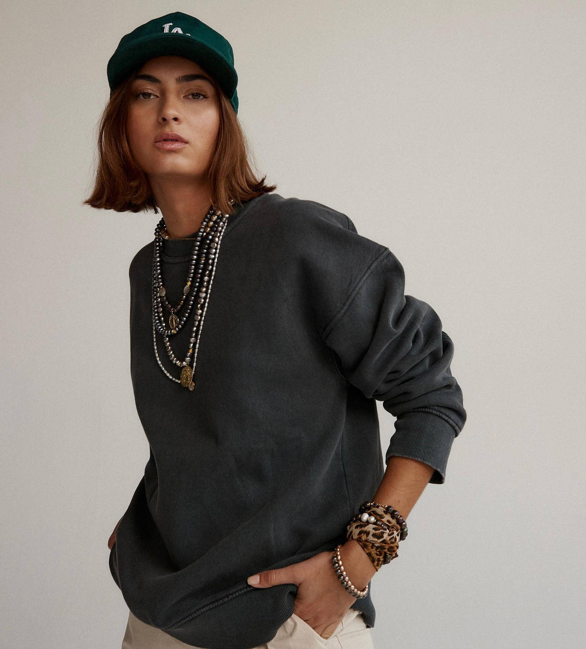 A model wearing a washed charcoal gray crewneck sweatshirt, layered with multiple silver and gold necklaces, a green baseball cap, and leopard print bracelets, set against a plain light gray background.