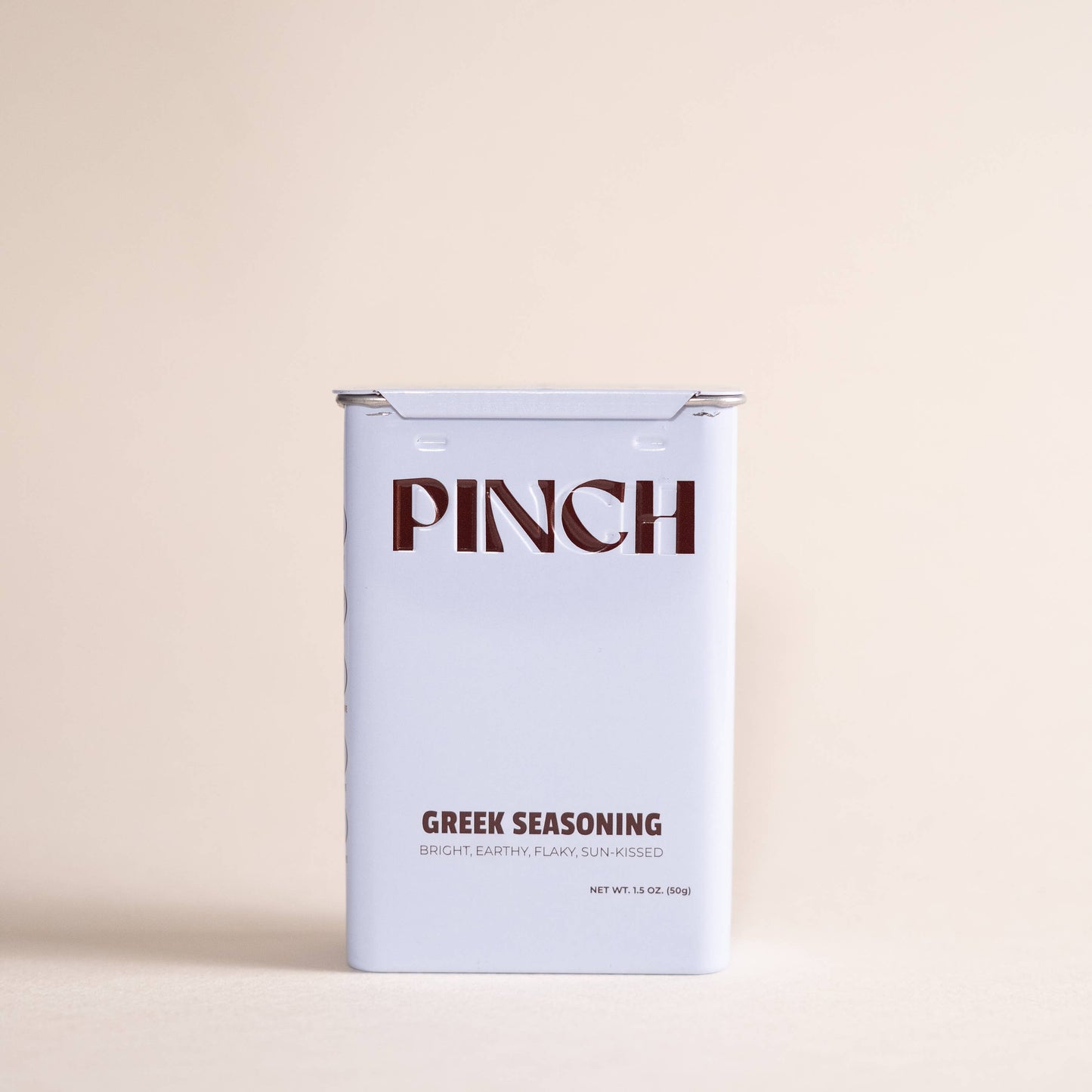 A rectangular tin of Pinch Greek Seasoning on a solid, warm beige background. The tin is white with dark brown text reading 'PINCH' and 'GREEK SEASONING'.