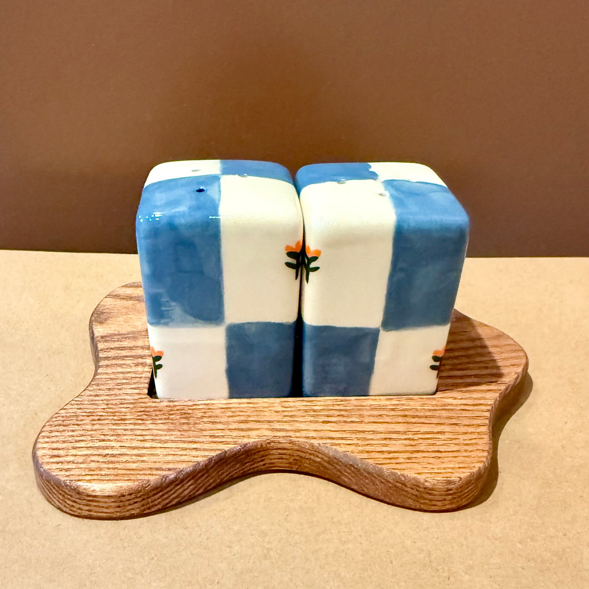 A pair of salt and pepper shakers with a blue and white checkered pattern, resting on a small, wavy wooden stand against a plain brown background.