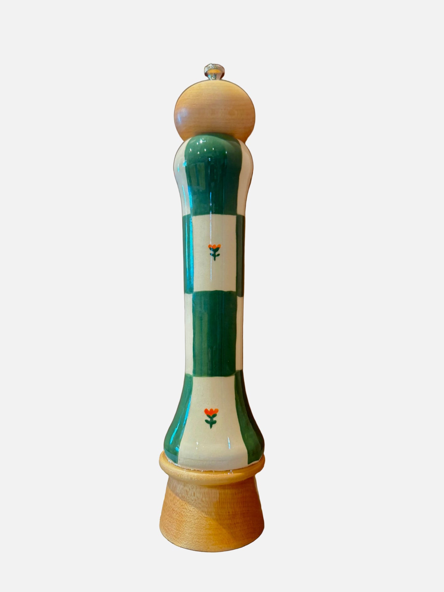 A decorative ceramic pepper mill with a green and white checkered body, small orange tulip details, a light wood top, and a light wood base, set on a marble-patterned surface against a warm brown background.