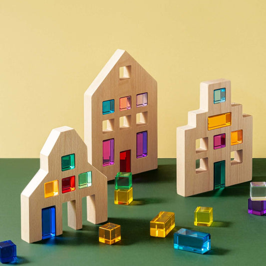 Colorful transparent blocks in front of wooden house models on a green surface with a yellow background