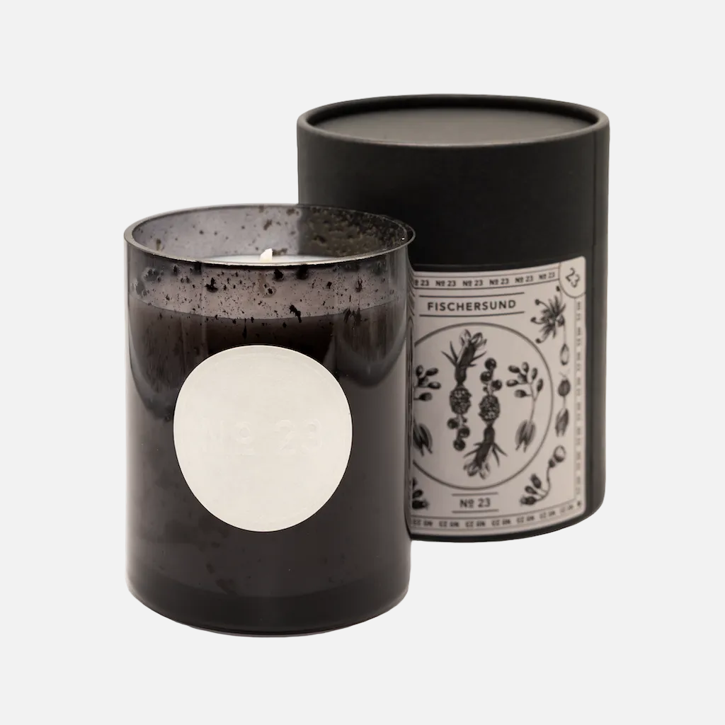 Black candle with decorative label next to its black cylindrical container on a white background