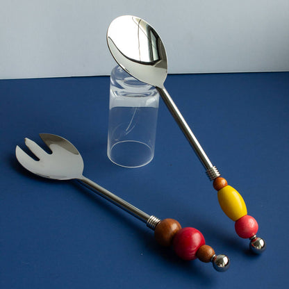 A set of stainless steel salad serving utensils, including a fork and a spoon, with colorful wooden bead handles in red, brown, yellow, and silver, arranged on a blue surface with a clear glass tumbler in the background.