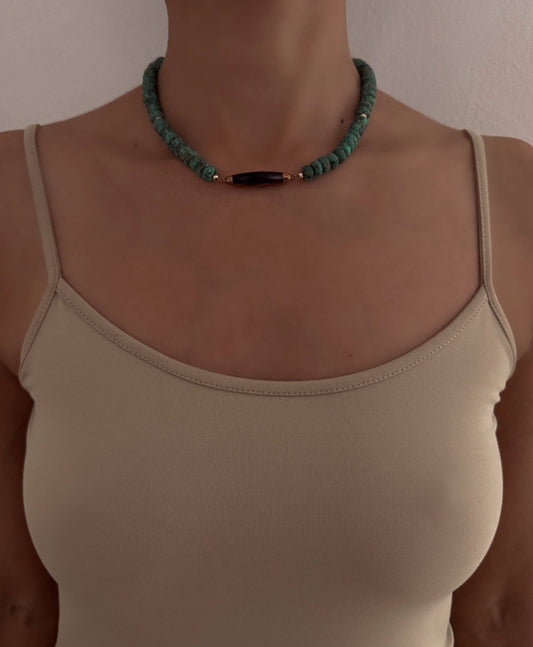 A close-up of a person wearing a turquoise beaded necklace with a central dark brown wooden bead and small gold spacer rings, paired with a beige spaghetti-strap top.