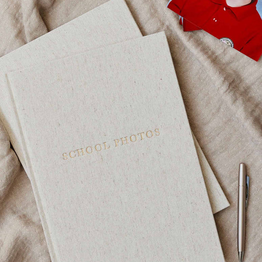 A linen-textured photo album titled 'SCHOOL PHOTOS' in gold foil, styled on a beige fabric background with a child's photo and a pen nearby.