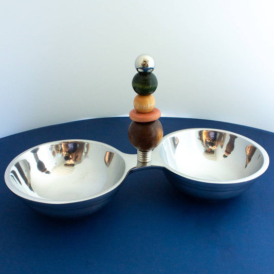 A modern two-bowl serving tray with a polished silver metal base and a central decorative column featuring stacked wooden and glass beads, set against a dark blue surface and light gray background.