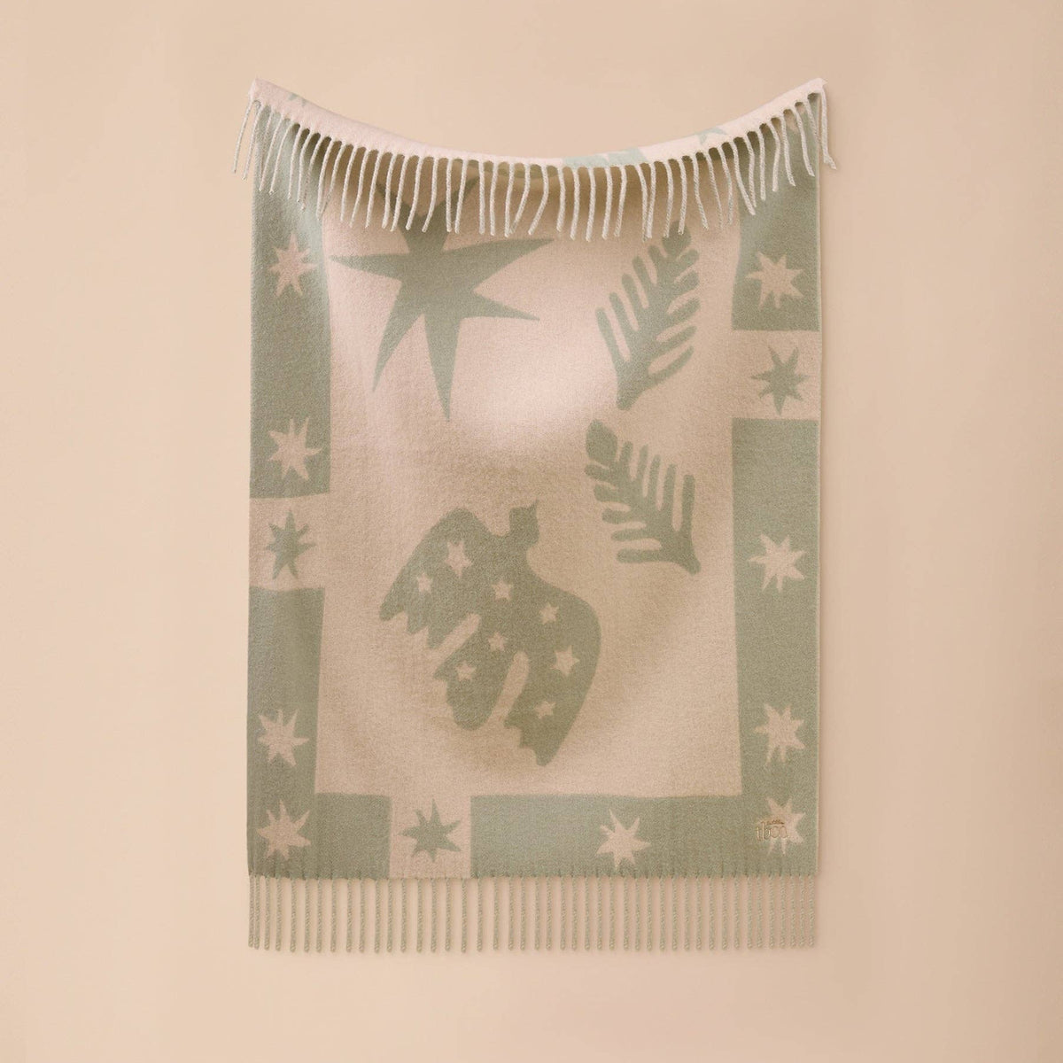 A light beige woven throw blanket with sage green patterns of stars, leaves, and a central bird silhouette, featuring white fringe on the top and bottom, displayed against a solid beige wall.