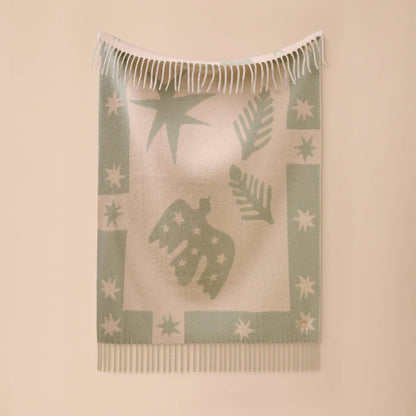 A light beige woven throw blanket with sage green patterns of stars, leaves, and a central bird silhouette, featuring white fringe on the top and bottom, displayed against a solid beige wall.