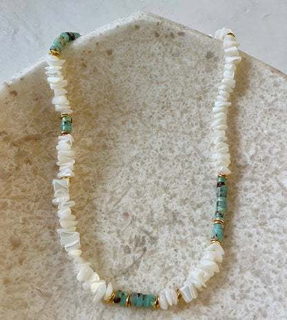 A beaded necklace made of white shell chips with small turquoise-blue beads and gold spacer rings, displayed on a textured beige stone surface.
