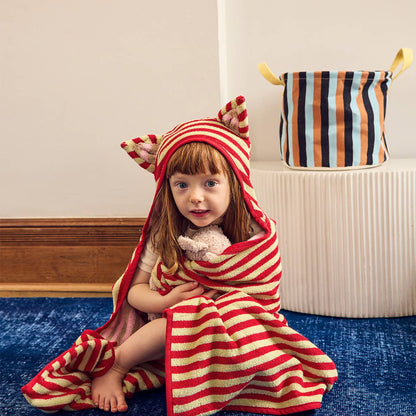 A young child sits on a blue carpet indoors, wrapped in a red and cream striped hooded towel with cat-ear details. A matching striped storage basket is visible on a white pedestal in the background.