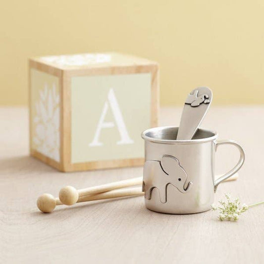 Metal mug with elephant design, wooden drumsticks, and spoon on a light surface.