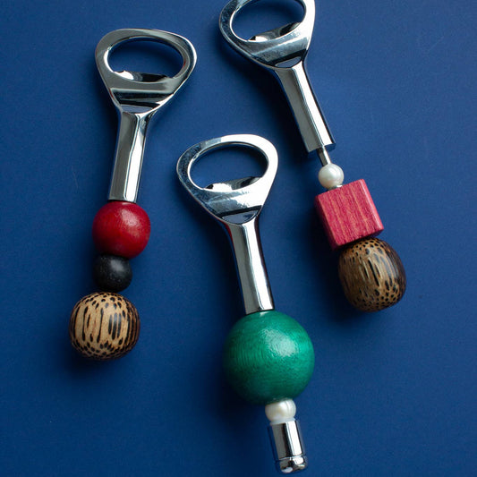 Three bottle opener keychains with colorful wooden beads and a pink block, displayed on a solid blue background.