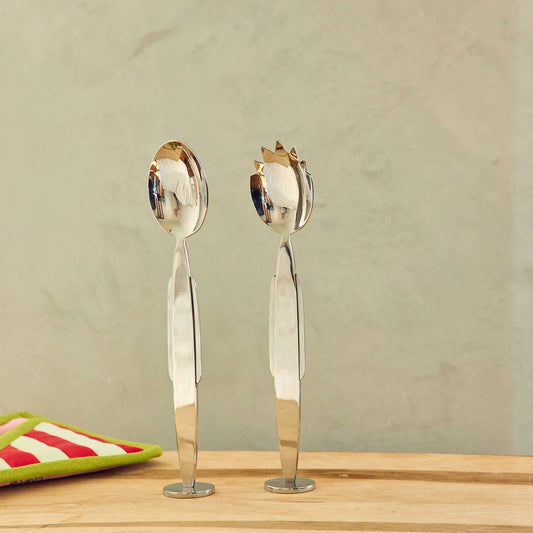 Two stainless steel salad spoons with decorative gold-toned handles, standing upright on a wooden surface against a textured beige wall.
