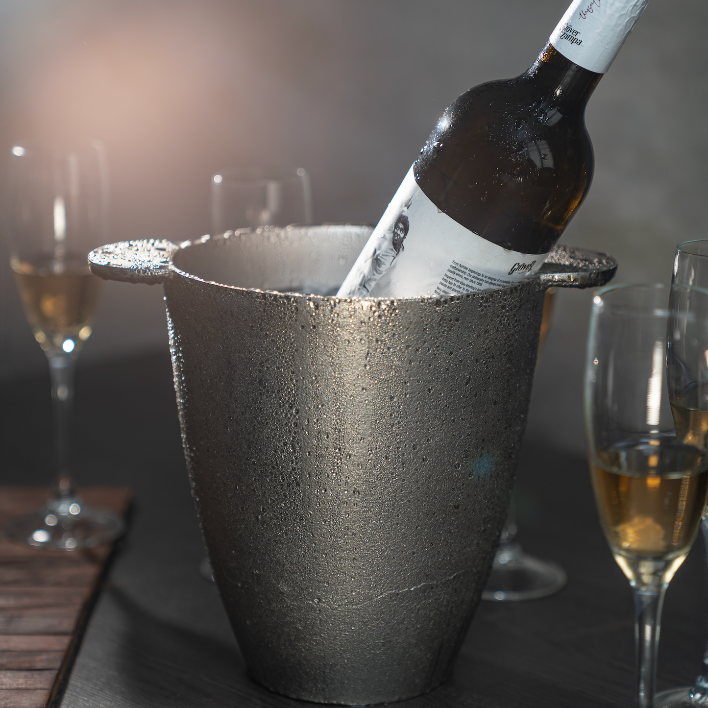 Wine bottle in an ice bucket with glasses around on a dark surface