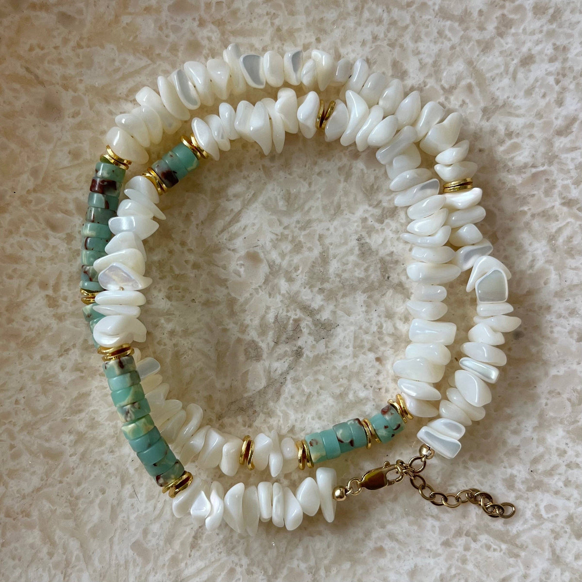 A beaded stretch bracelet made of white shell-like disc beads, turquoise-blue spacer beads, and small gold-tone metal spacers, displayed on a textured beige surface.