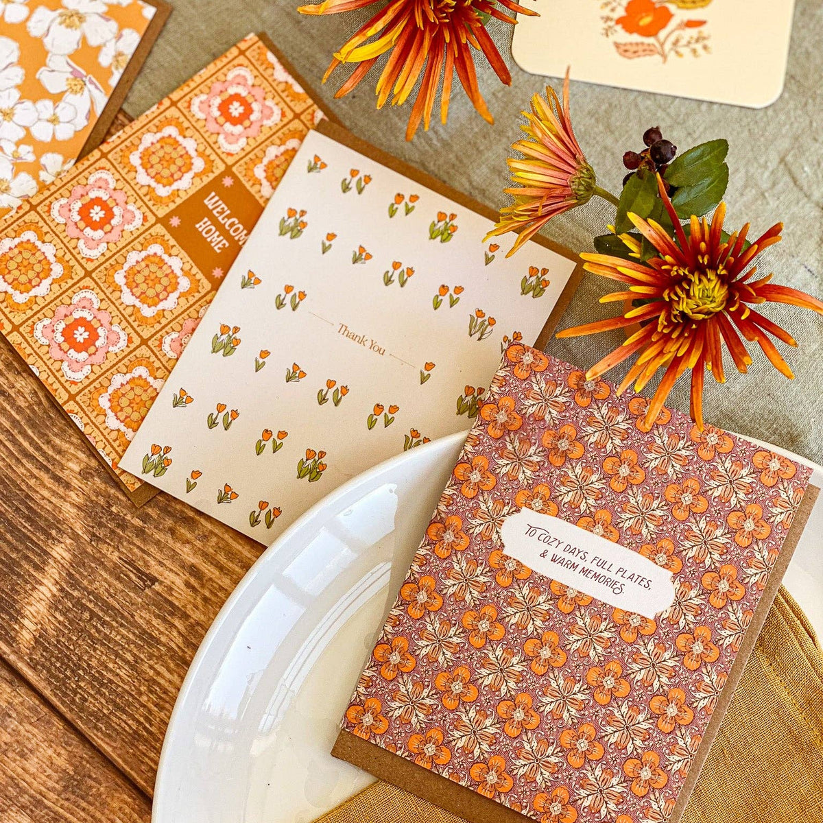 A styled flat-lay of autumn-themed stationery, including patterned cards, a small notebook or journal with a floral cover and a label reading 'IT'S YOUR LAND, NOT MY APPLE & PEACH MANGOES,' decorative flowers, a white plate, and a rustic wood surface.