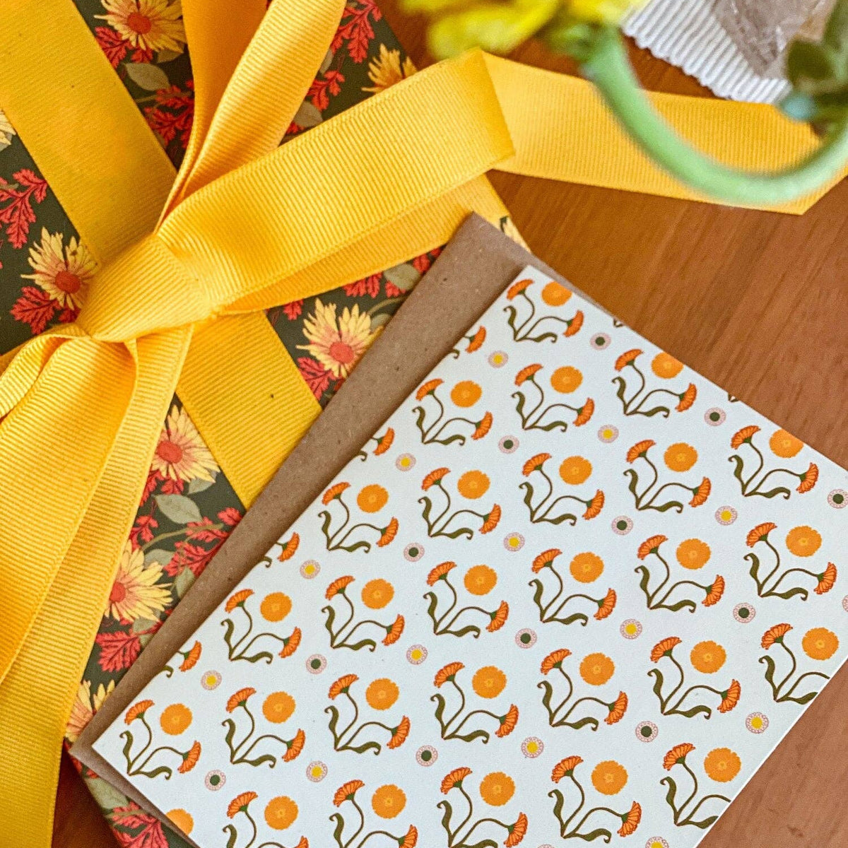 A close-up of gift wrapping paper and ribbon on a wooden surface. One wrapped package has a dark green floral pattern with yellow and orange flowers and a wide yellow ribbon bow, while another package features a white background with a repeating orange flower and brown stem pattern.