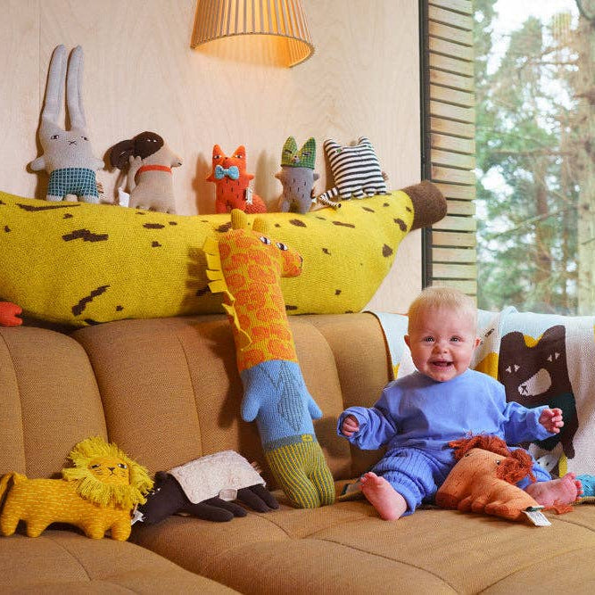 Child playing with colorful stuffed animals on a brown couch in a room with a lamp and window.