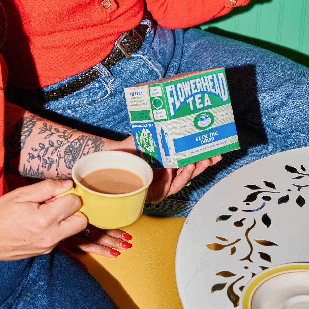 A person in a red shirt and blue jeans holds a box of Flowerhead Tea while sitting at a table with yellow teacups and saucers against a green wall.