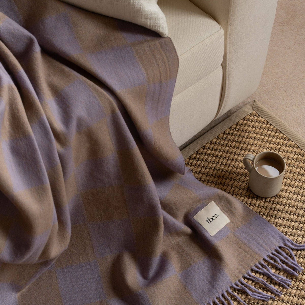 Plaid blanket draped over a sofa with a mug and small container on a textured rug.