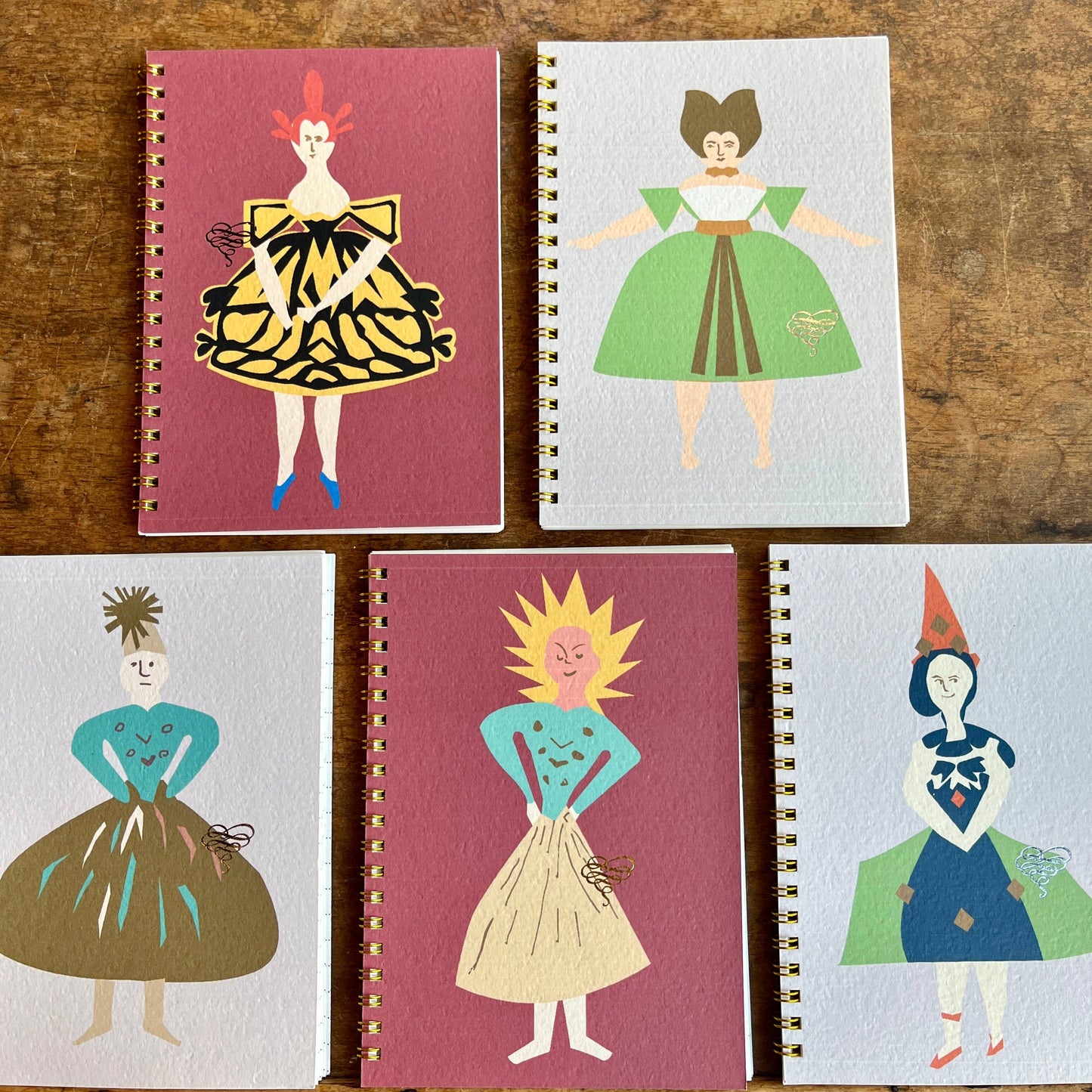 Five notebooks with illustrated covers of people in colorful outfits on a wooden surface.