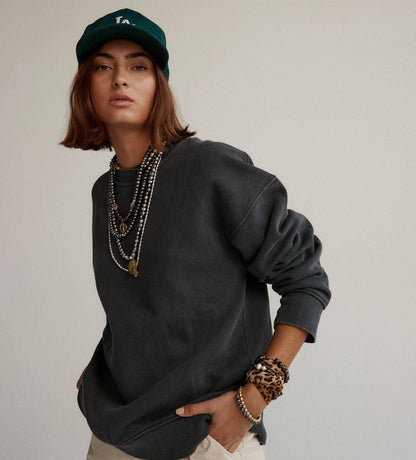 A model wearing a washed charcoal gray crewneck sweatshirt, layered with multiple silver and gold necklaces, a green baseball cap, and leopard print bracelets, set against a plain light gray background.