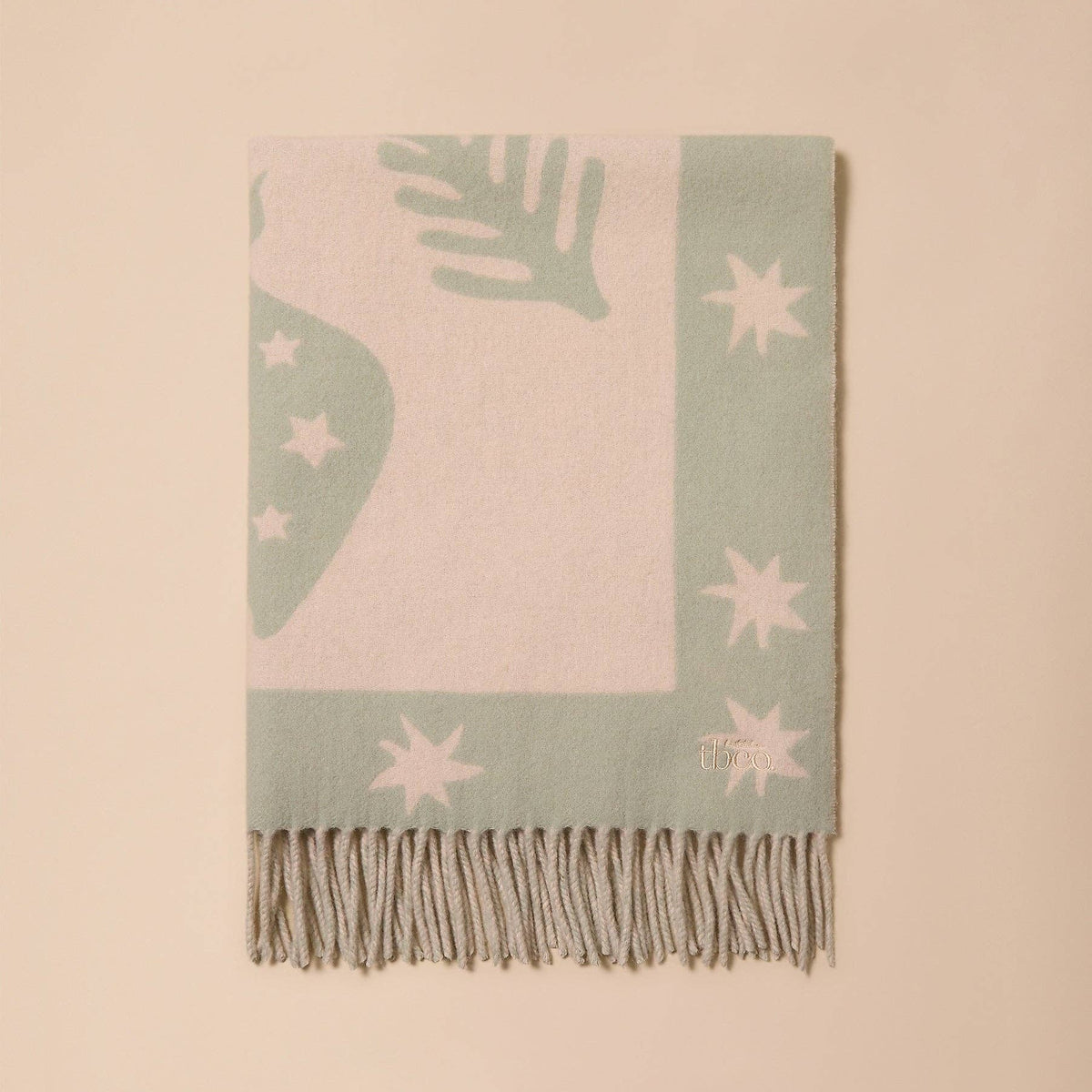 A rectangular woven throw blanket with fringe, featuring a pale pink center and a sage green border with white star and leaf motifs, displayed flat against a solid beige background.