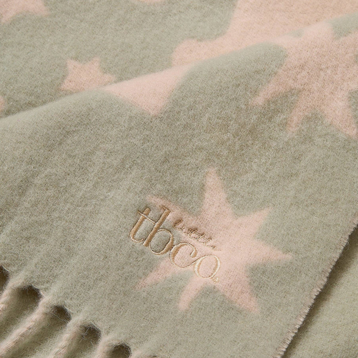 A close-up of a soft fleece throw blanket with a pale sage green and light pink star pattern, featuring a gold embroidered 'tbco' logo and fringed edges.