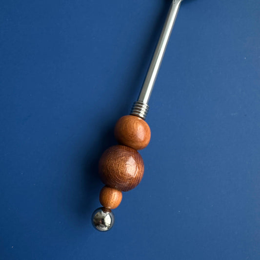A stainless steel spoon with a decorative handle featuring three stacked wooden beads in warm brown tones and a small metallic ball at the end, set against a solid blue background.