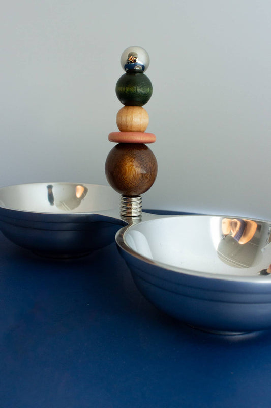 A decorative metal and wood bowl set with a central stacked-berry handle featuring silver, dark green, light wood, terracotta, and dark brown spherical accents, shown on a blue surface against a gray background.
