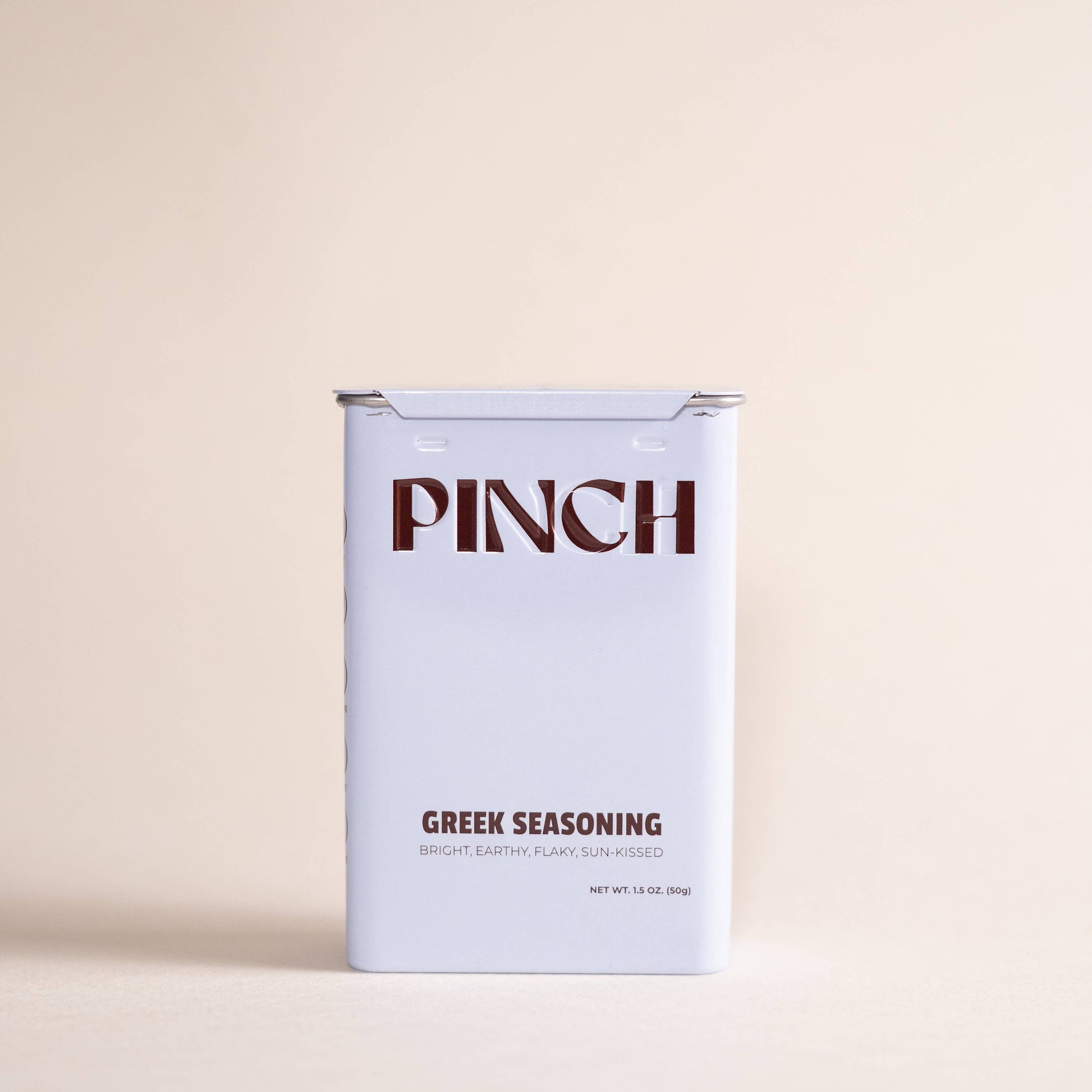 A rectangular tin of Pinch Greek Seasoning on a solid, warm beige background. The tin is white with dark brown text reading 'PINCH' and 'GREEK SEASONING'.