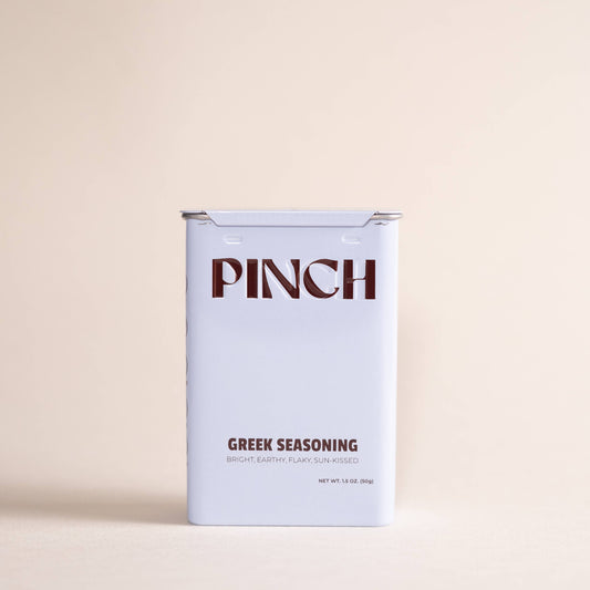 A rectangular tin of Pinch Greek Seasoning on a solid, warm beige background. The tin is white with dark brown text reading 'PINCH' and 'GREEK SEASONING'.
