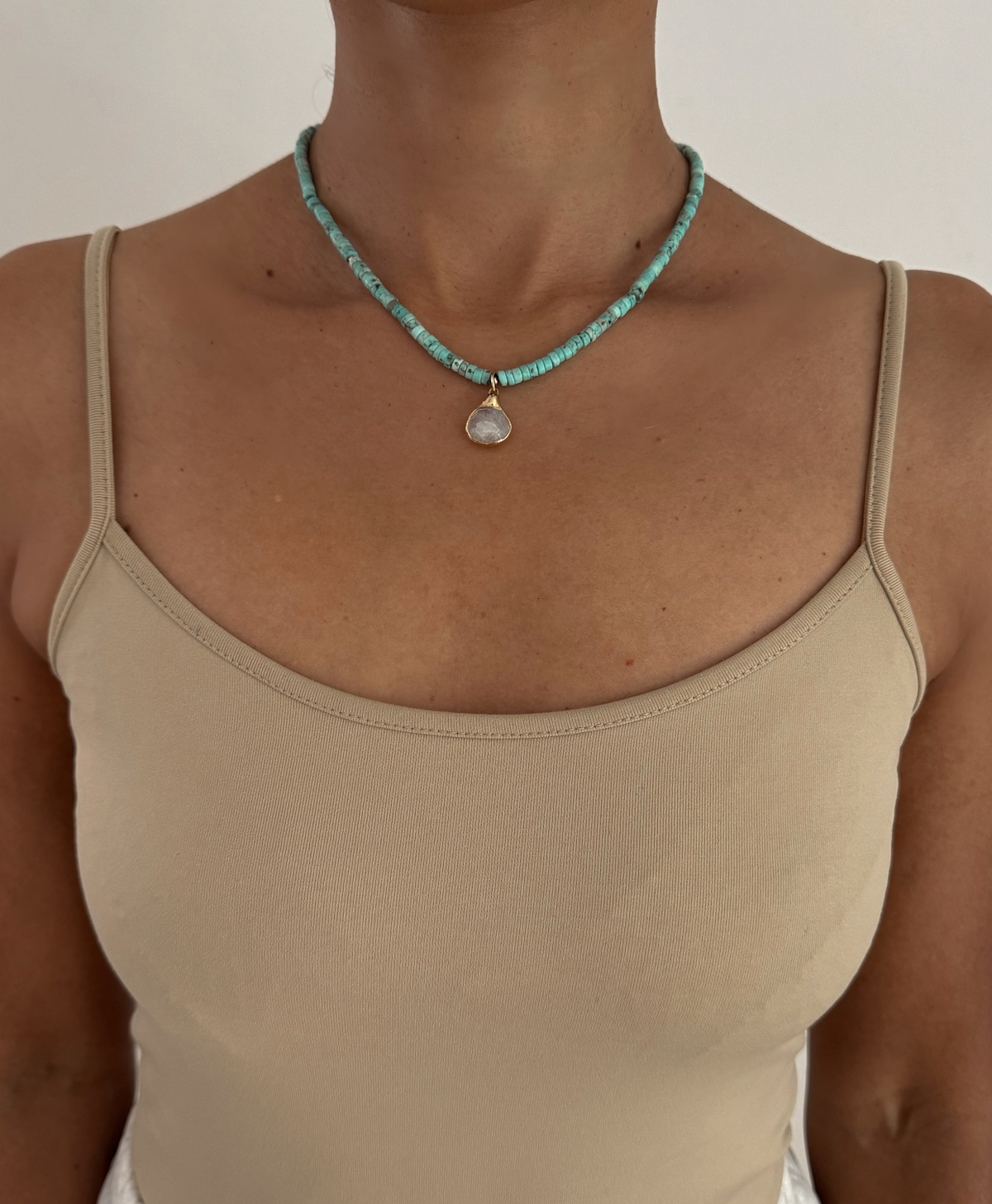 A close-up of a model wearing a turquoise beaded necklace with a teardrop-shaped white pendant, styled with a beige spaghetti-strap top against a plain off-white background.