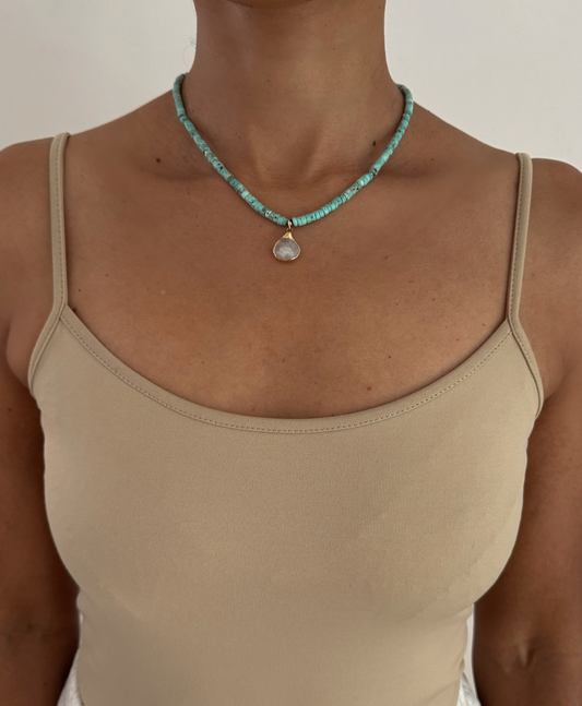 A close-up of a model wearing a turquoise beaded necklace with a teardrop-shaped white pendant, styled with a beige spaghetti-strap top against a plain off-white background.