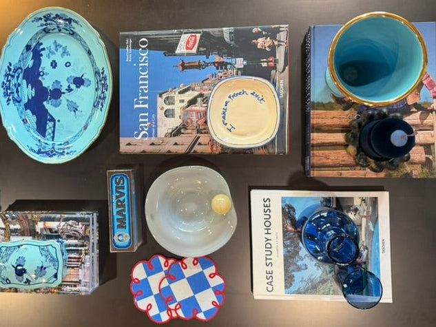A top-down view of a dark wooden coffee table displaying a collection of blue and white ceramic tableware, including plates, cups, and a teacup, alongside a Marvis box and a Case Study Houses brochure, set on a patterned area rug.
