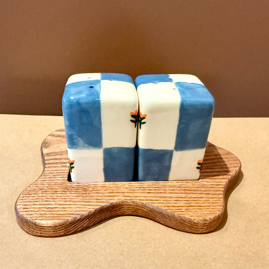 A pair of salt and pepper shakers with a blue and white checkered pattern, resting on a small, wavy wooden stand against a plain brown background.