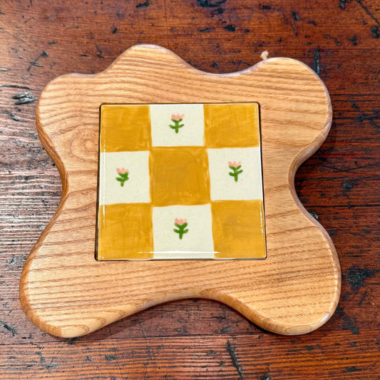 A scalloped wooden tray or coaster with a central square inset featuring a yellow and white checkerboard pattern with small pink flowers, shown on a dark wood tabletop.