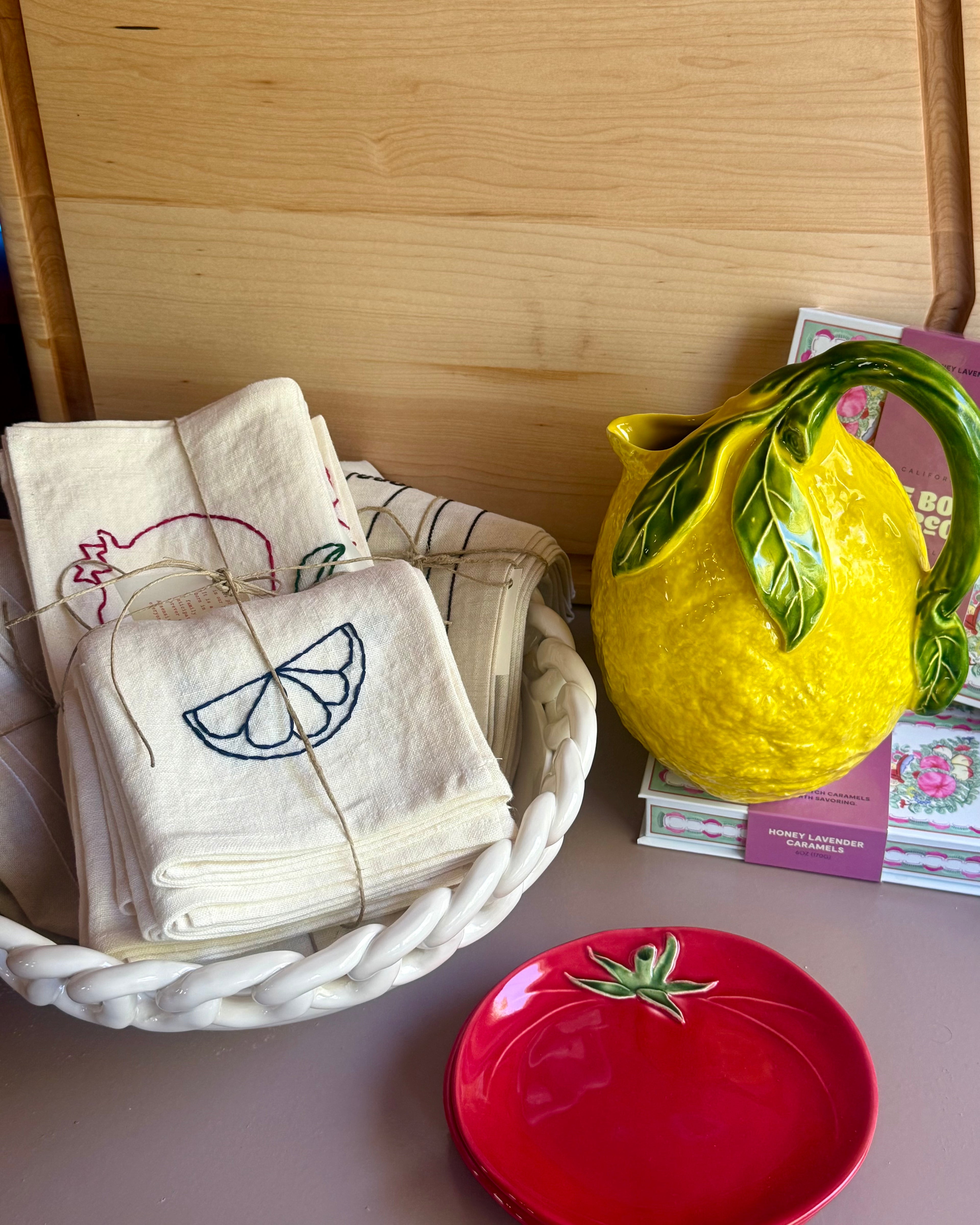 Bowl with embroidered towels, yellow lemon-shaped pitcher, and red dish on a wooden surface.