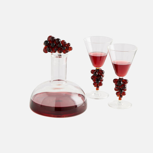 Wine decanter with red wine and two glasses, each with a decorative stem of red grapes on a white background.