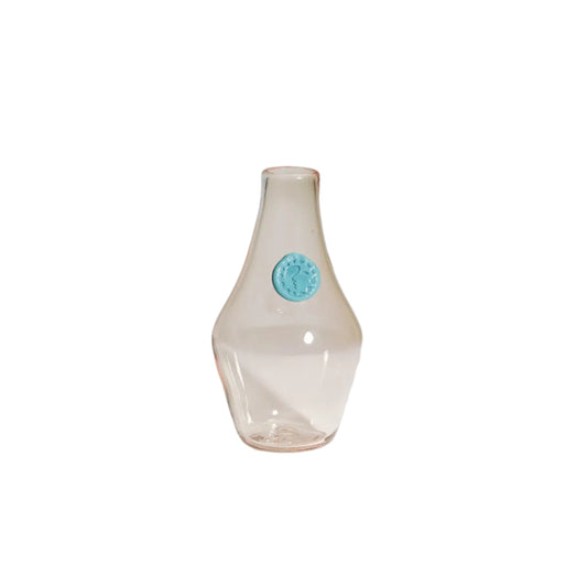 A small clear glass vase with a pale pink tint and a round turquoise blue emblem on the front, set against a light gray studio background.