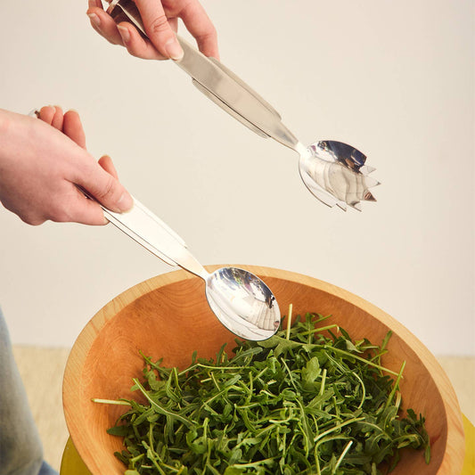 A pair of stainless steel salad servers with white handles being used to toss green salad in a wooden bowl, held by hands against a plain light background.