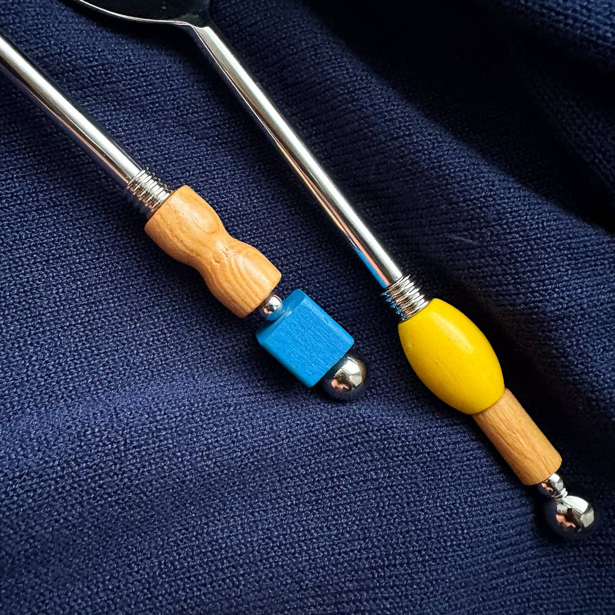 Two metal utensils with wooden handles and colorful end accents, placed on a dark navy textured fabric background.