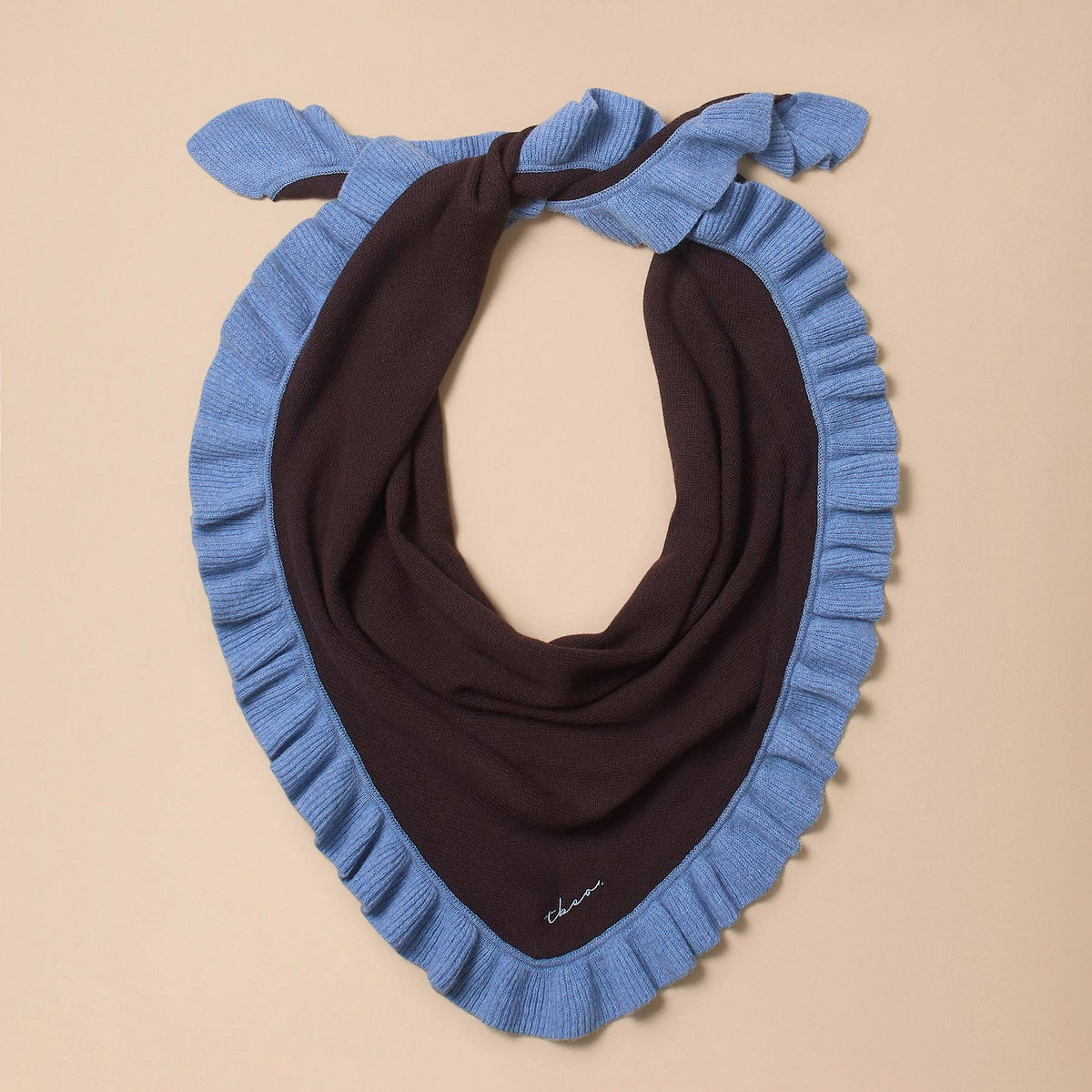 A knit neck warmer scarf with a light blue fringed border and a dark brown center, knotted at the top, displayed on a solid beige background.