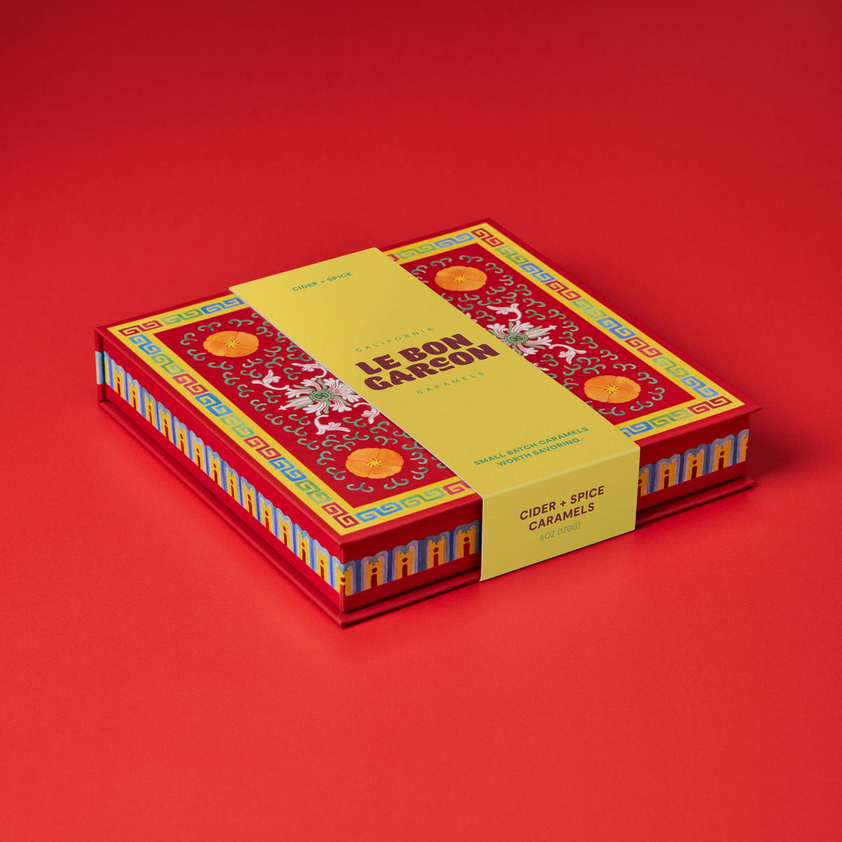 A boxed product labeled 'LE BON GARSON' and 'GOURD & SPICE CARAMELS' sits on a solid red studio background. The box is red with ornate gold and multicolor decorative patterns, and a yellow paper band wraps around the middle.