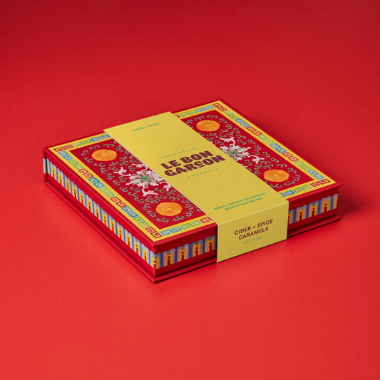 A boxed product labeled 'LE BON GARSON' and 'GOURD & SPICE CARAMELS' sits on a solid red studio background. The box is red with ornate gold and multicolor decorative patterns, and a yellow paper band wraps around the middle.