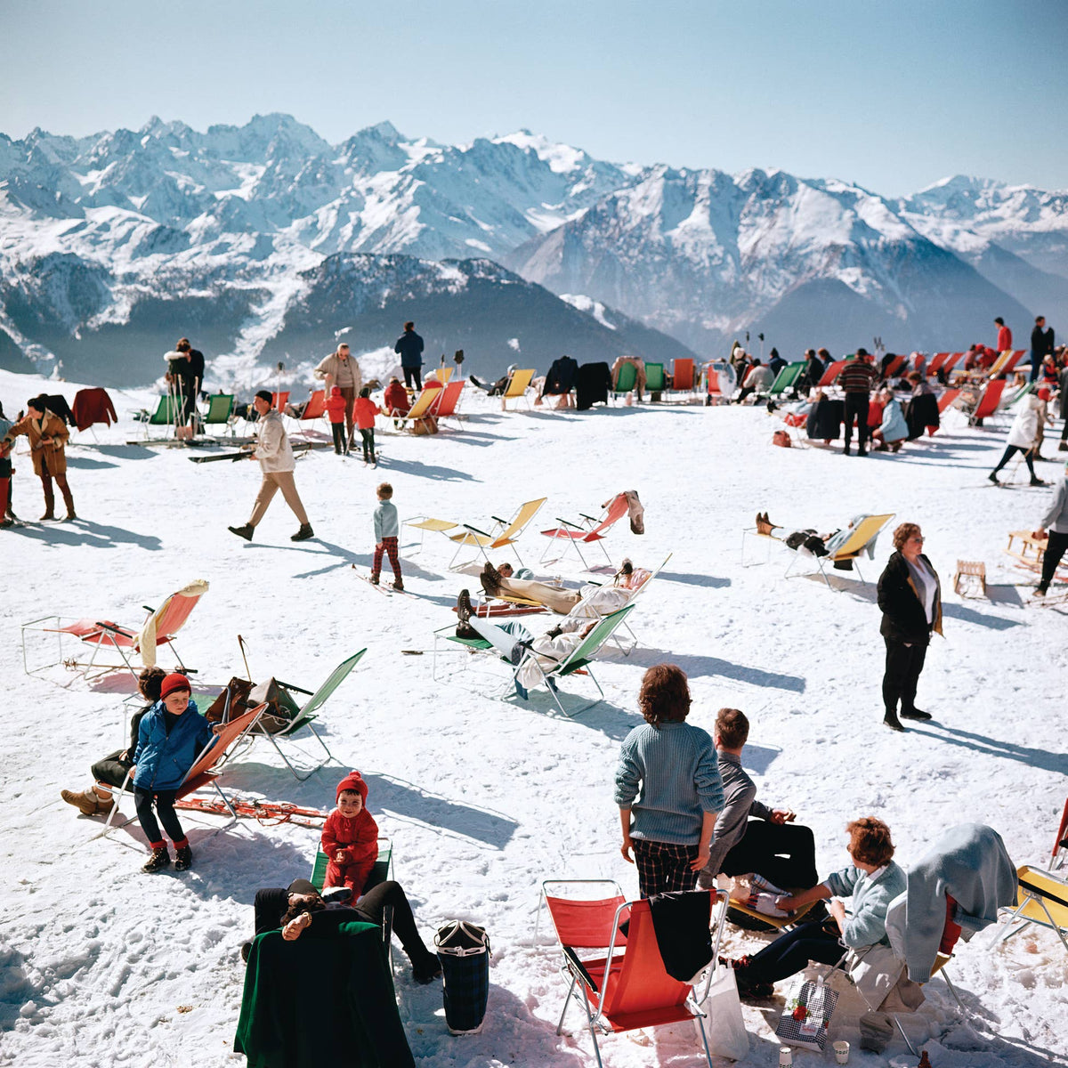 People enjoying a sunny day on a snowy mountain with colorful chairs and mountains in the background.